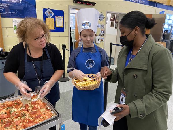 Two adults and a student serve pizza from a rolling cart